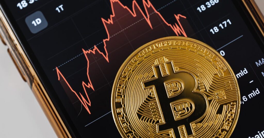 A close-up of a gold Bitcoin coin placed on a smartphone displaying a stock market chart.